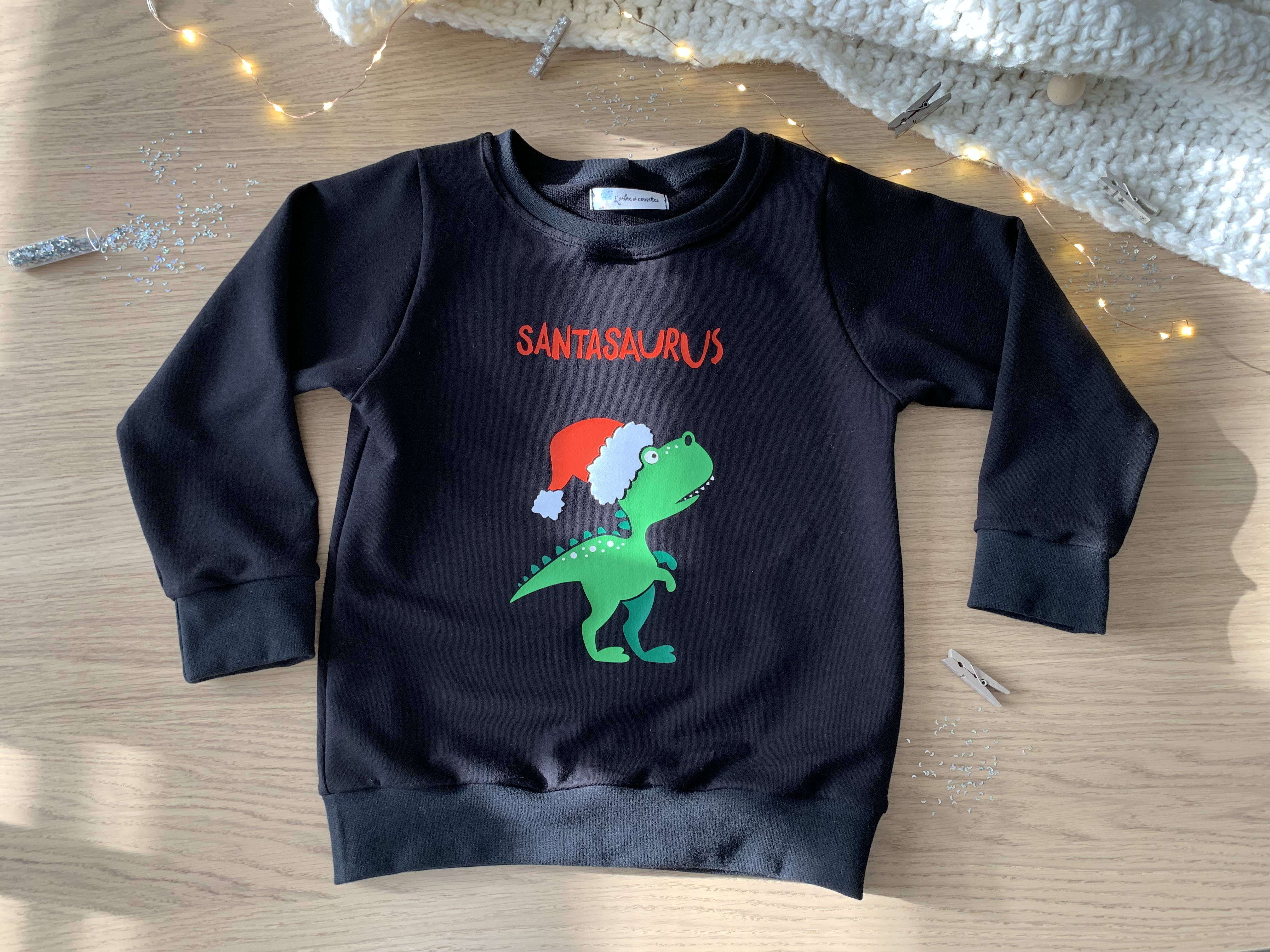 Pull de Noël enfant cousu en sweat noir avec motif dinosaure, posé près d’une guirlande